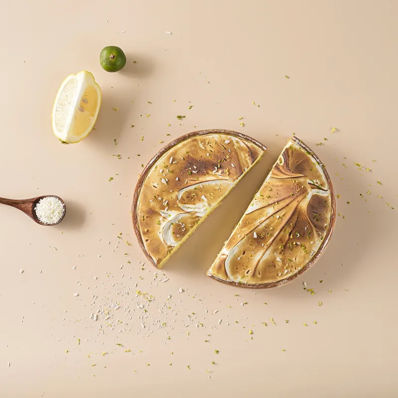 Overhead view of a calamansi meringue tart cut in half on a warm cream surface, with a lemon wedge, a whole calamansi and scattered zest.