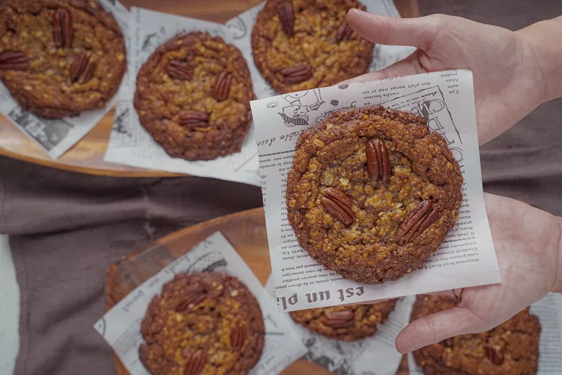Two hands holding a pecan cookie on printed paper above a tray of more cookies.