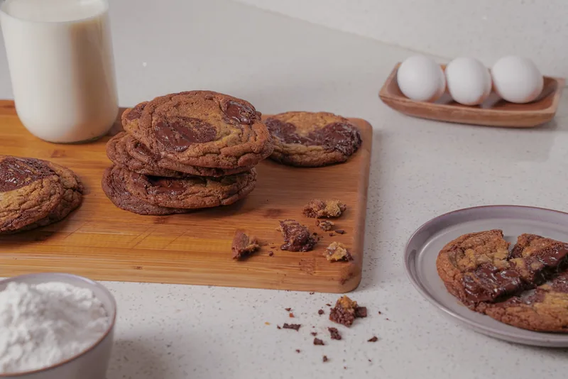Cookies stacked on a wooden board beside a glass of milk, three eggs and a bowl of flour.