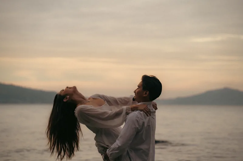 A man lifts his partner at the water's edge as the sun sets behind low hills.