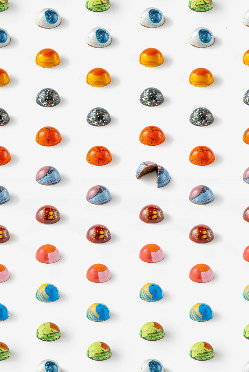 Rows of hand painted bonbons in every colour on white, one cracked open to show the filling.