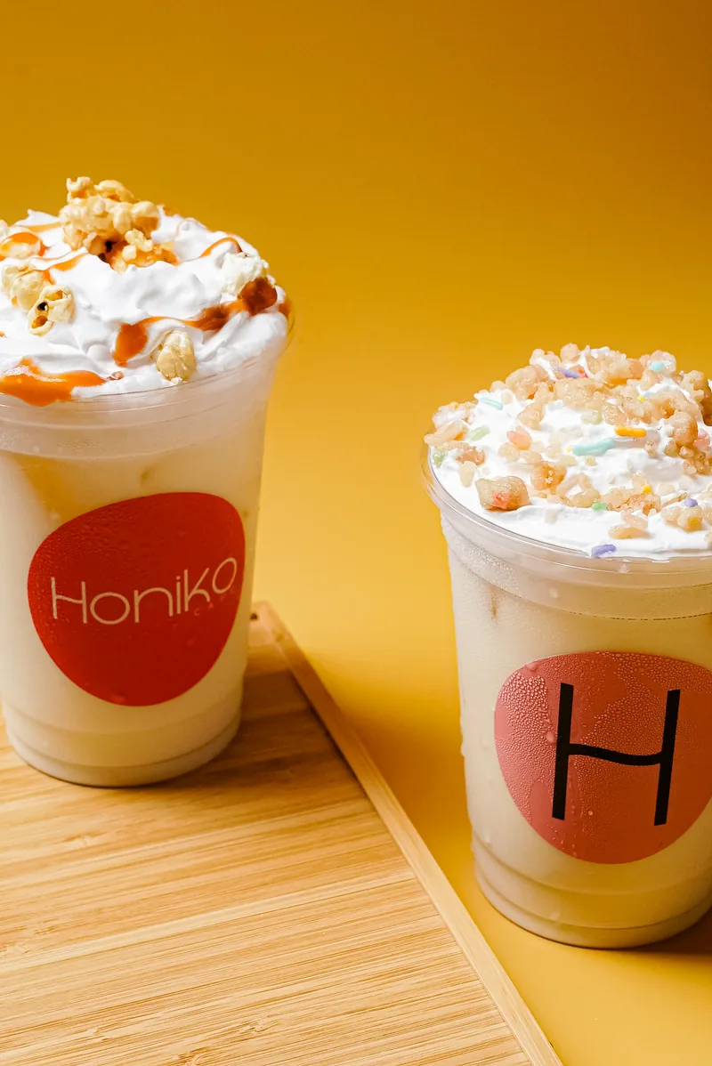 Two Honiko cream drinks on a wooden paddle against a yellow wall.