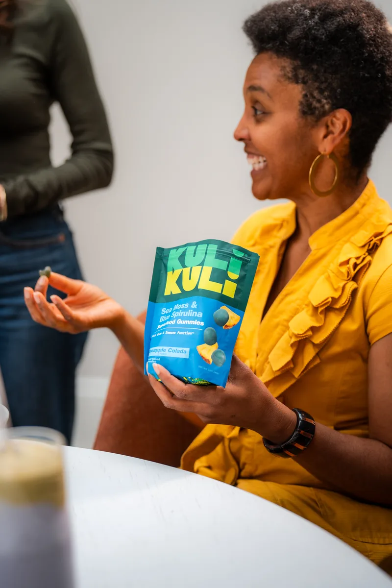 A woman in mustard holding a Kuli Kuli pouch toward the camera and smiling.