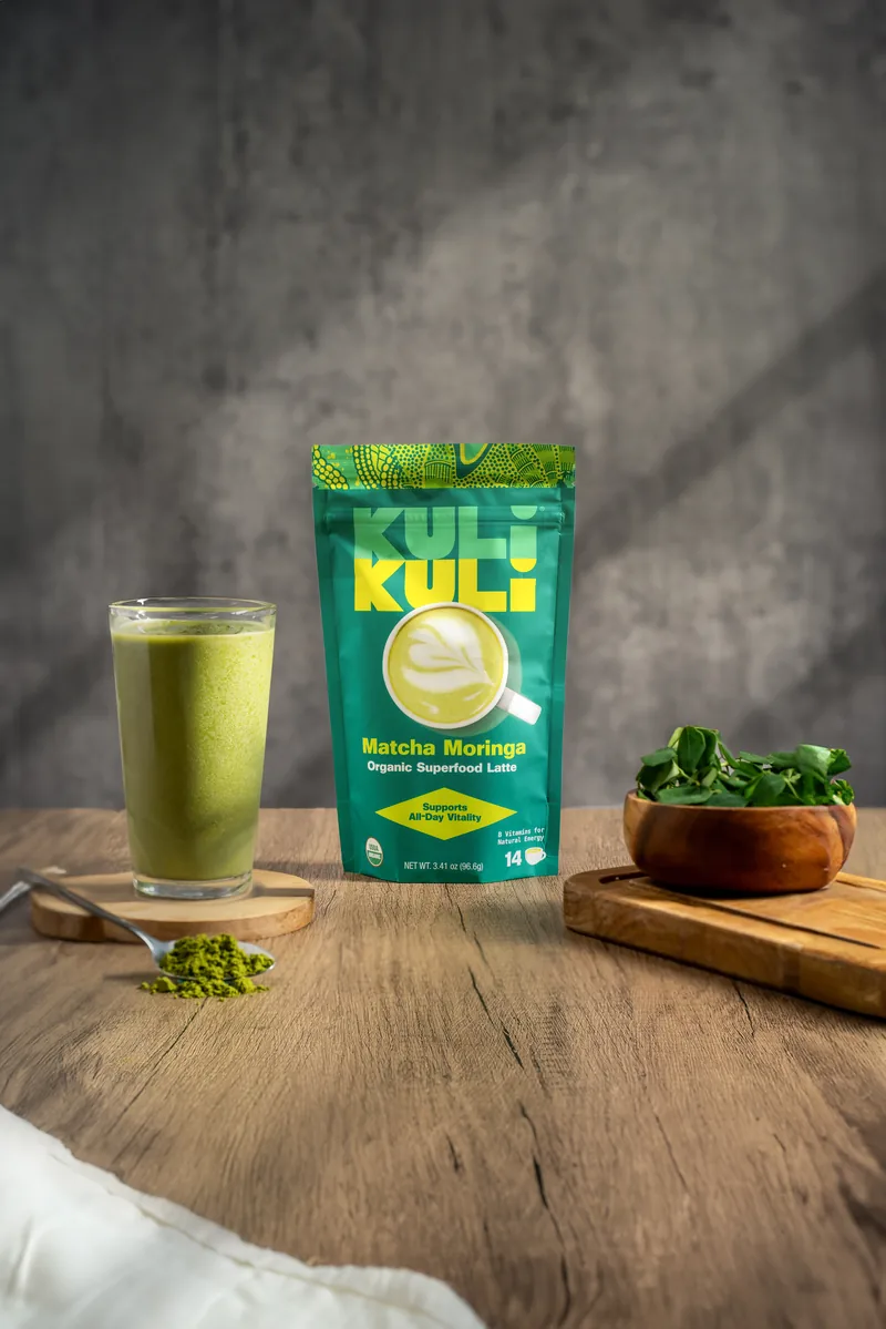A Kuli Kuli Matcha Moringa pouch standing beside a green latte and a bowl of moringa leaf on a wooden table.