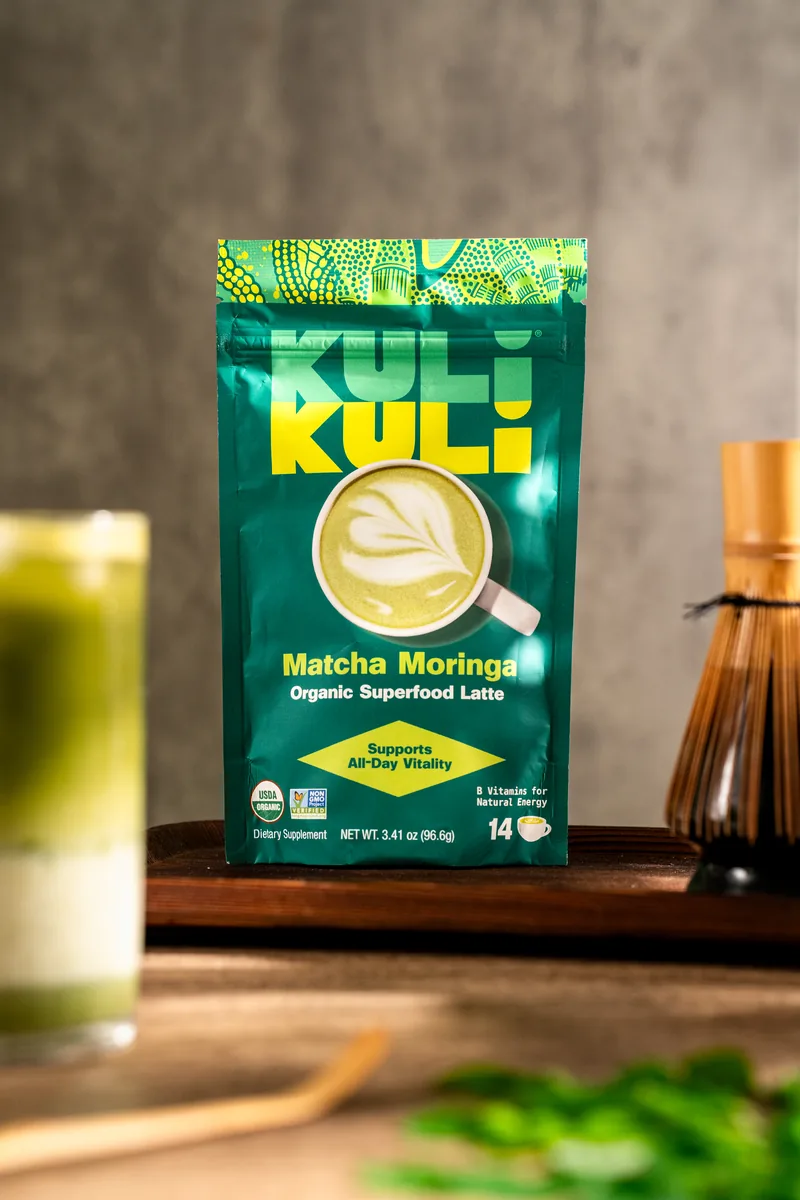 A Kuli Kuli Matcha Moringa pouch standing front on, a layered matcha latte soft behind it.