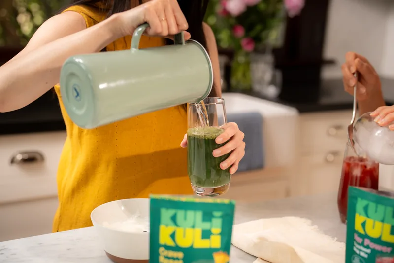 A green superfood smoothie being poured from a jug into a glass in a bright kitchen.