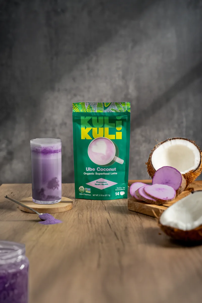 A Kuli Kuli Ube Coconut pouch with a violet iced latte, halved coconut and sliced ube beside it.