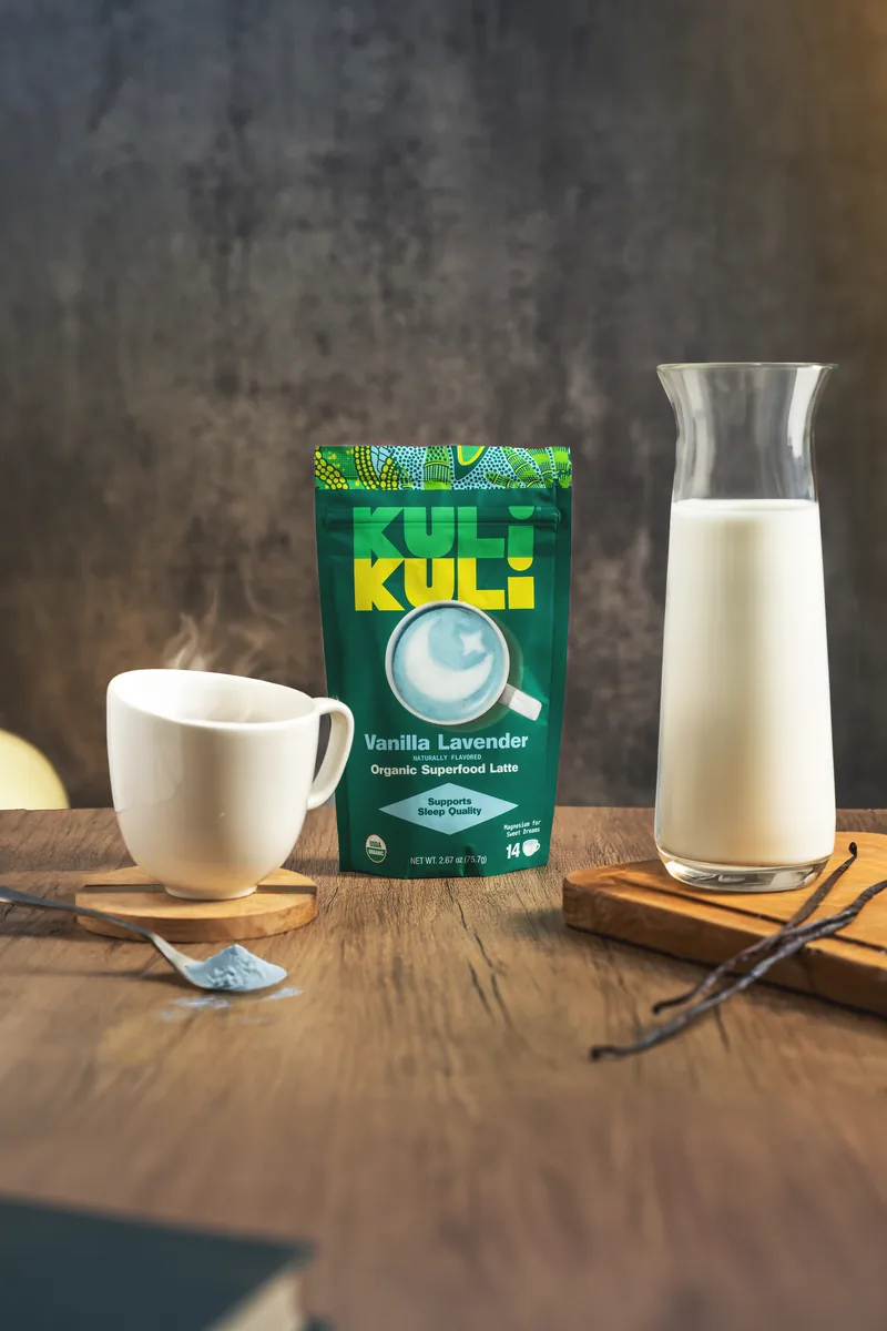 A Kuli Kuli Vanilla Lavender pouch beside a white mug, a carafe of milk and vanilla pods.