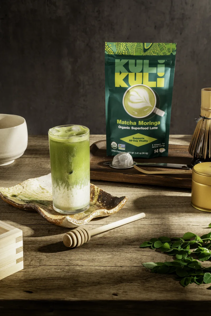 A layered matcha latte in a tall glass beside a bamboo whisk, a honey dipper and a Kuli Kuli pouch.