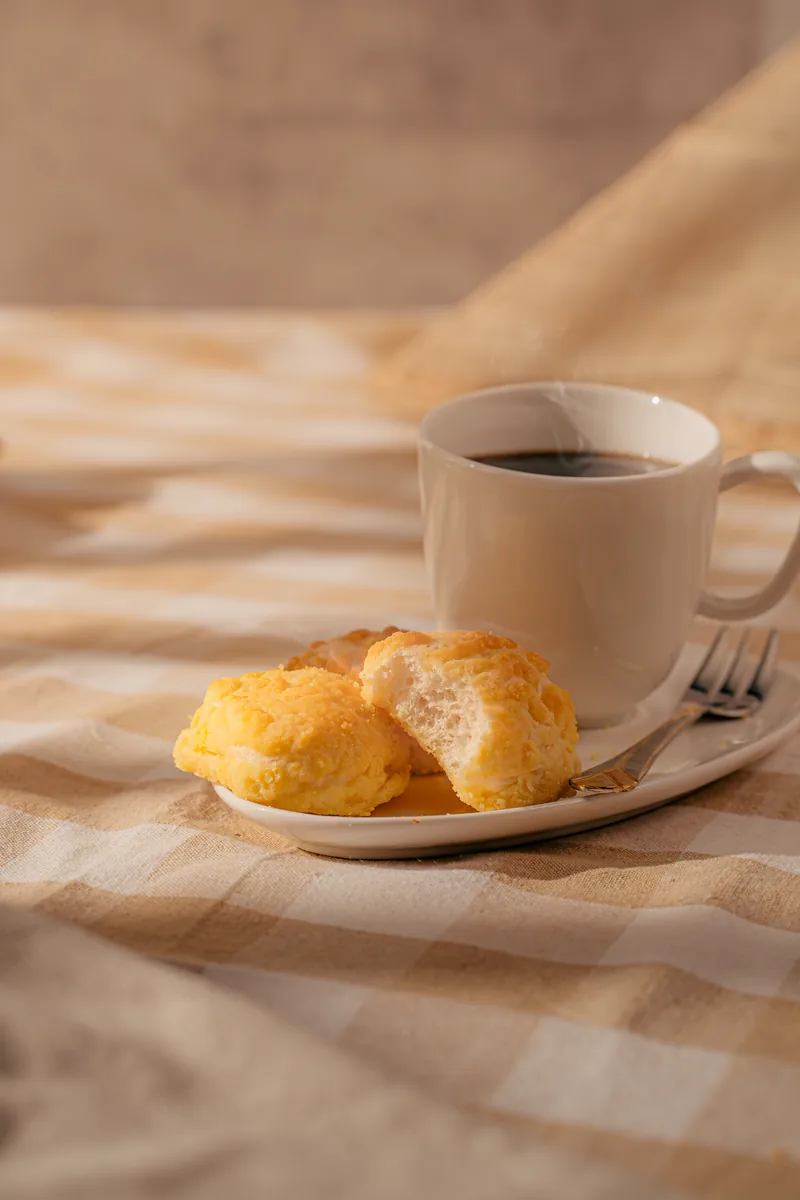Cheese bread on a saucer beside a cup of black coffee on a checked tablecloth.