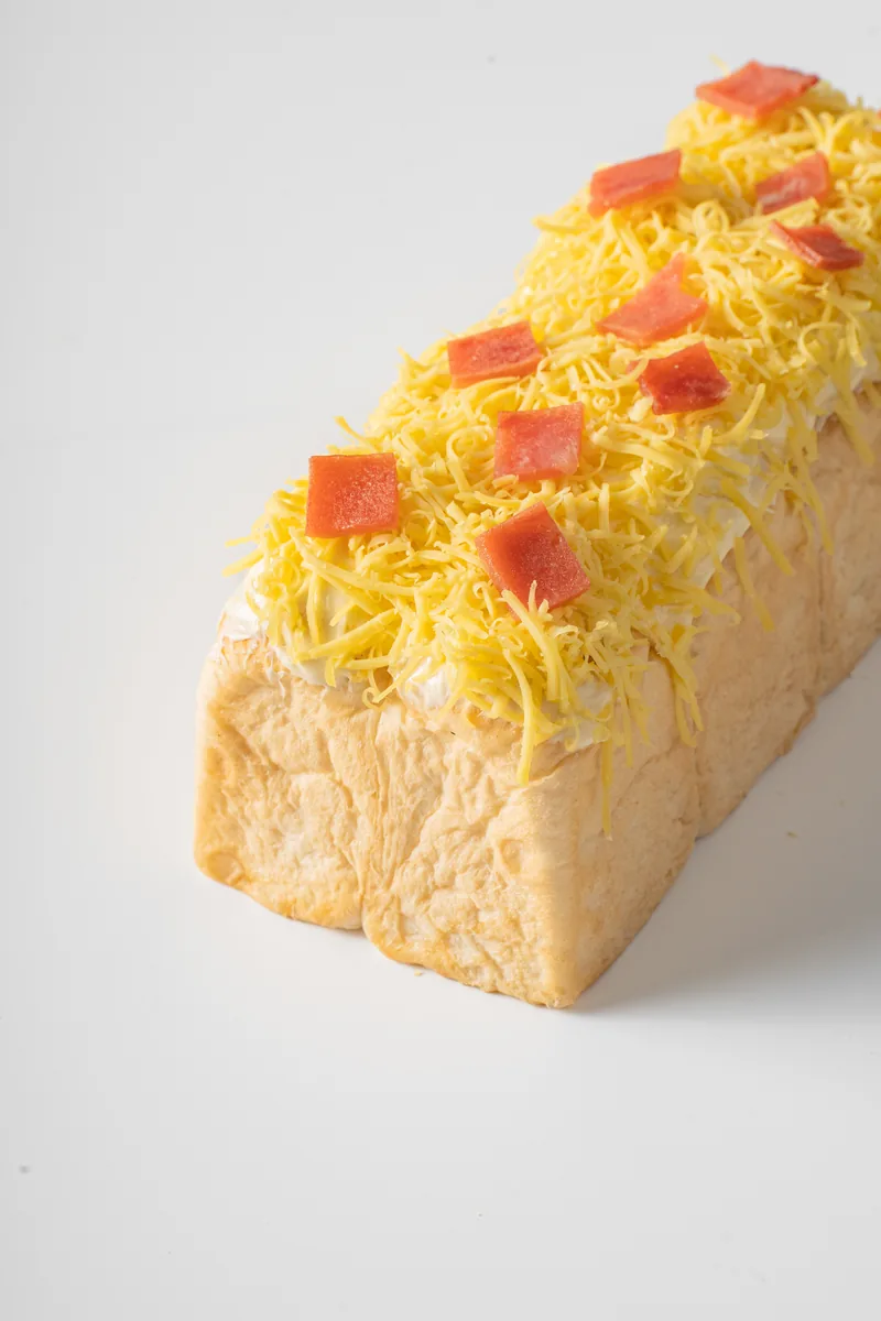 A long ensaymada loaf topped with cheese and red bits, on the diagonal.