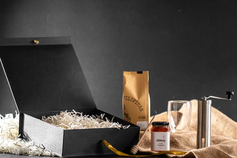 An open gift box with a kraft coffee bag, jar and hand grinder on burlap.