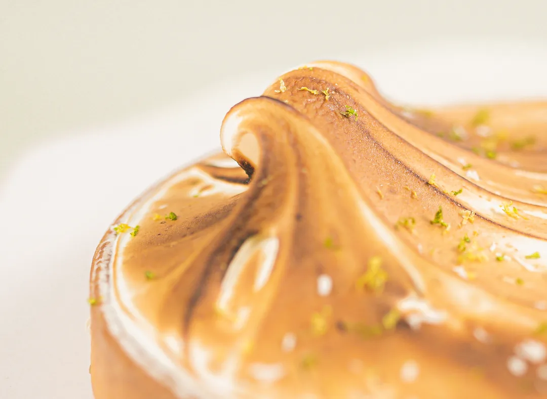 Extreme close-up of torched meringue, its glossy folds catching warm light with flecks of citrus zest.