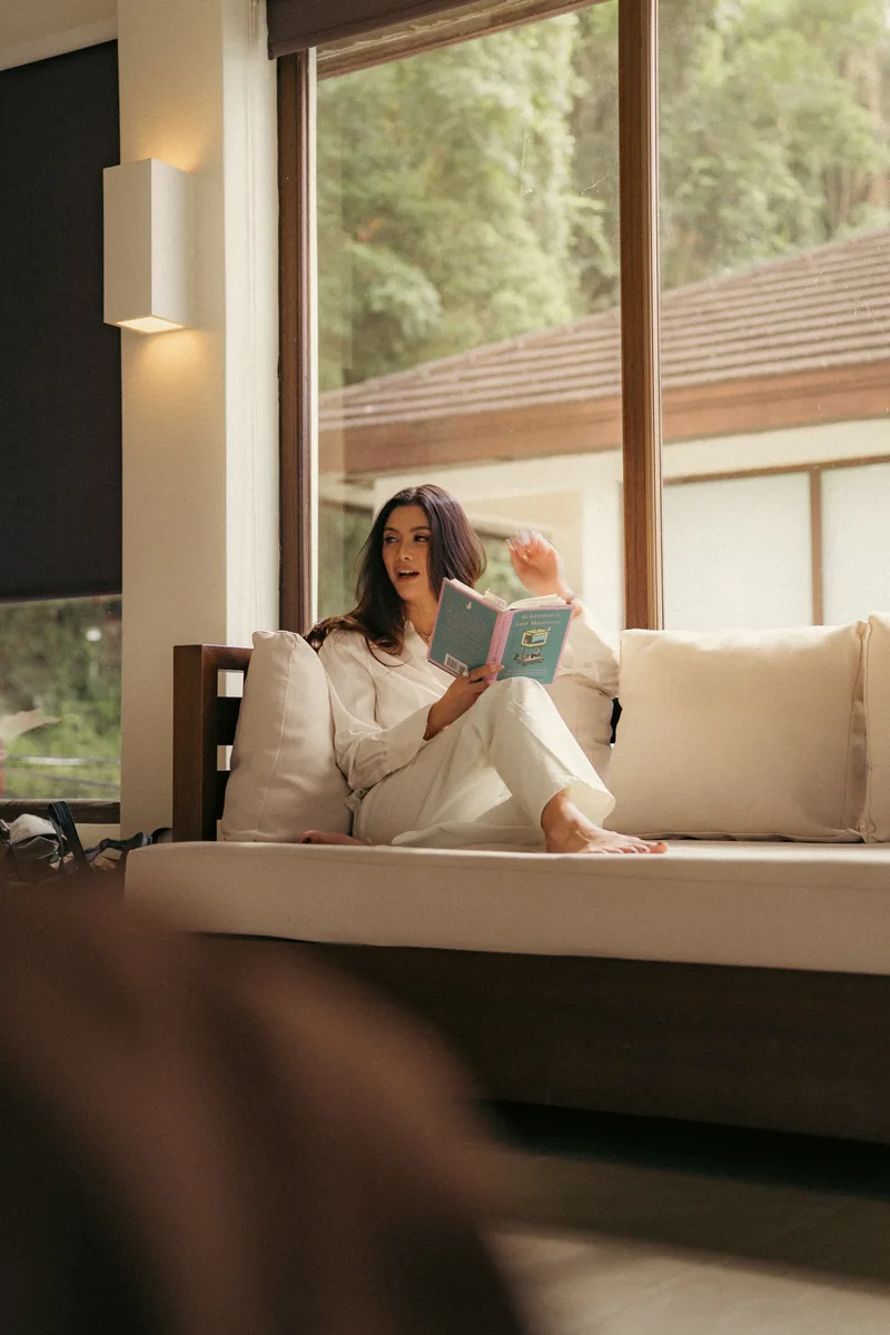 A woman reads on a pale sofa beside floor-to-ceiling windows opening onto trees.
