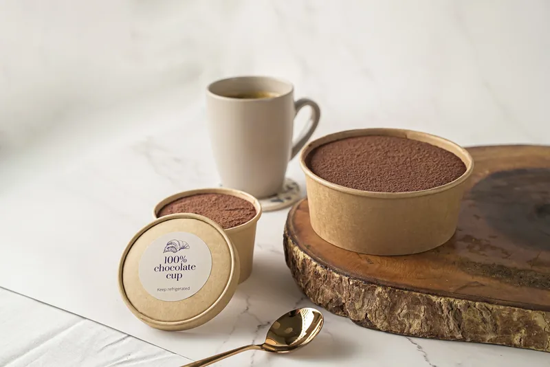A kraft tub of chocolate cup dessert dusted with cocoa, beside a mug on a wooden board.