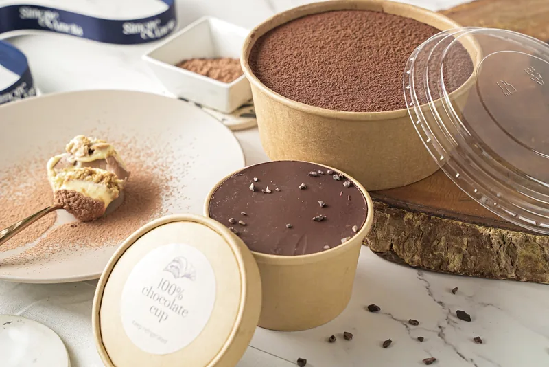 Kraft tubs of chocolate cup dessert with lids off on marble, cocoa powder and chocolate alongside.