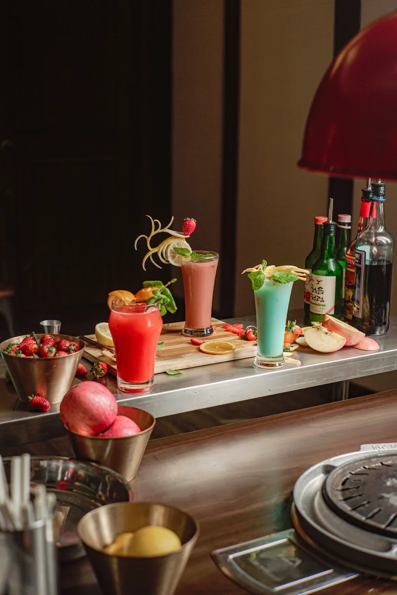 A bar pass lined with pink, red and mint coloured drinks, fresh fruit and bottles behind.