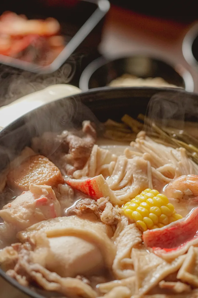 Steam rising off a shabu pot packed with mushrooms, tofu, corn and crab, shot close.