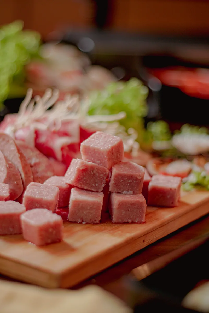 Cubes of marbled raw beef lined up on a wooden board with greens behind.