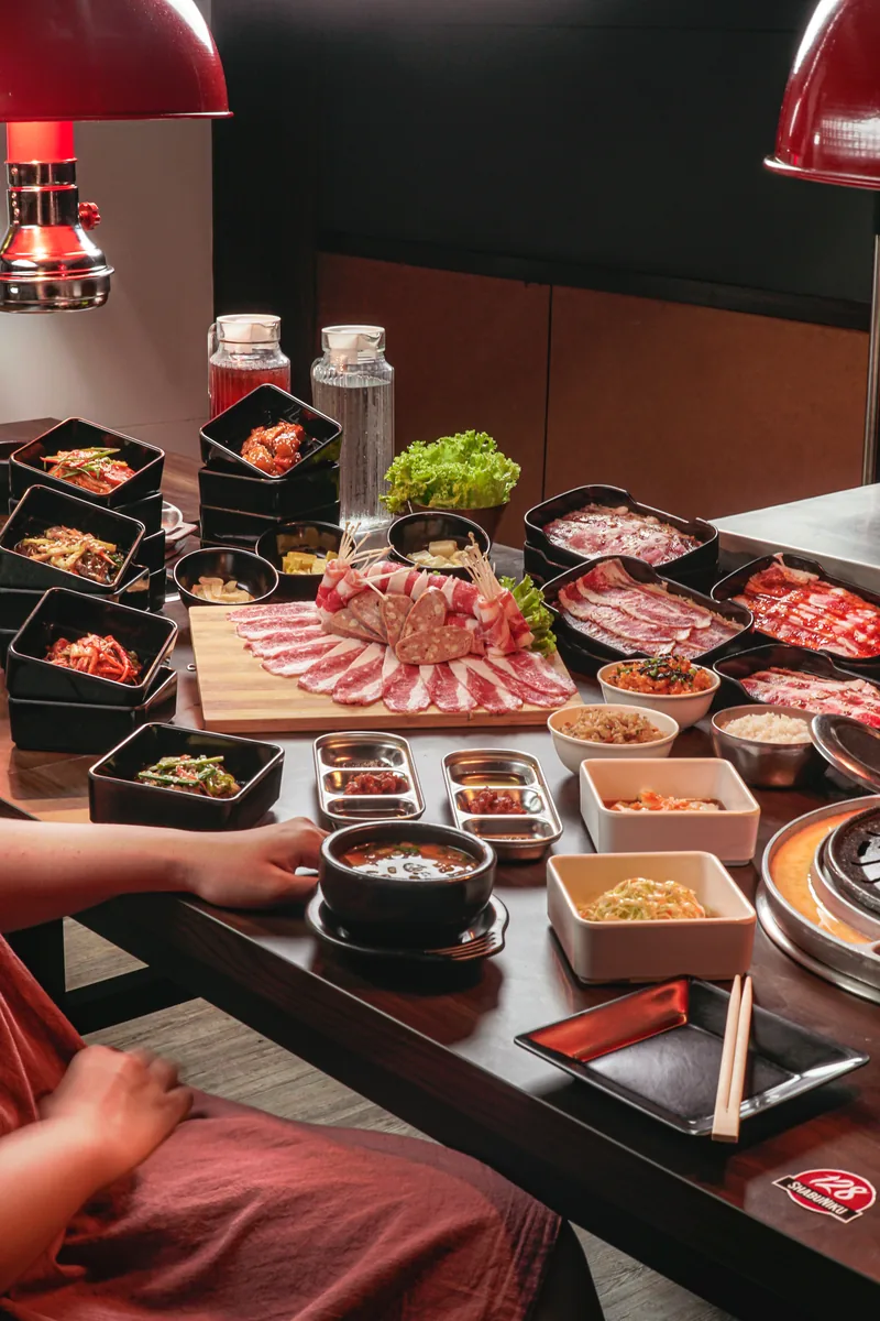 A long table under red lamps set with raw meat platters, banchan bowls and a shabu pot, a diner reaching in from the left.