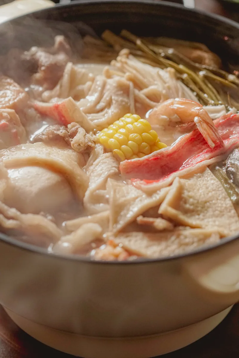 A steaming hotpot crowded with corn, fish balls and sliced pork.