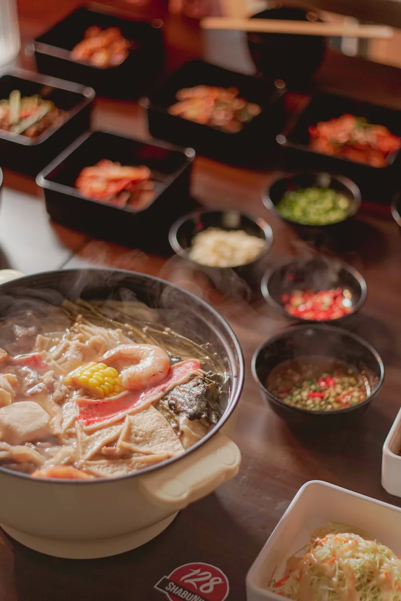 A shabu pot of broth and vegetables set on a dark table with side dishes and dipping sauces behind it.