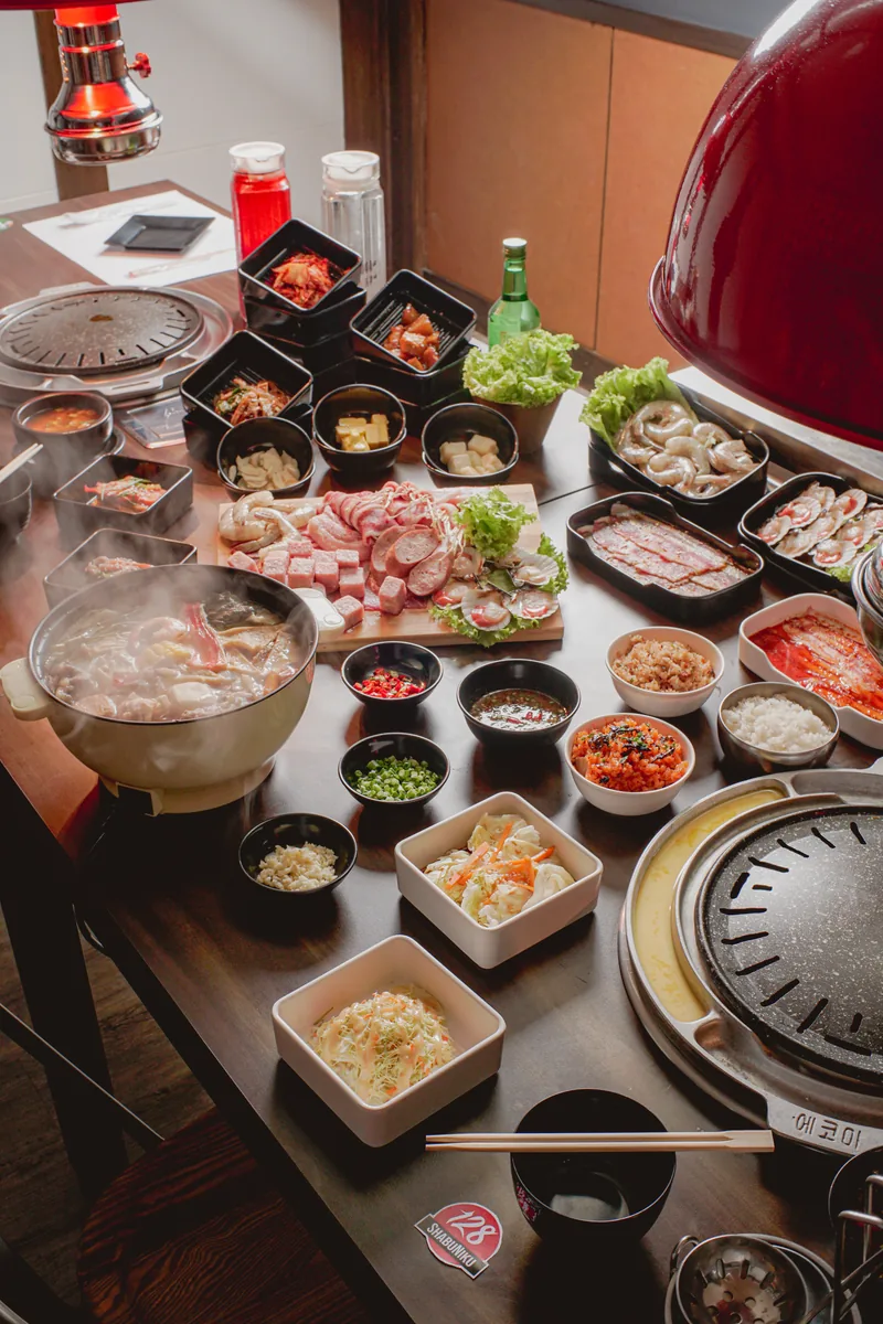 A vertical view down a dark table set with banchan, meat and a steaming pot.