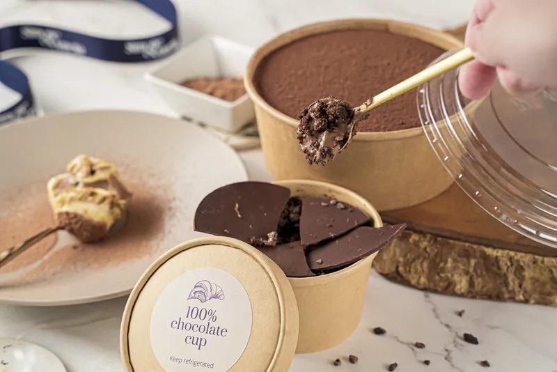 A spoon lifting a scoop from a chocolate cup in its kraft tub.